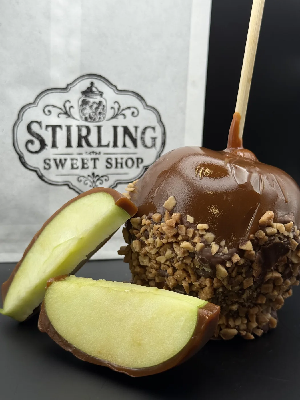 A nut-covered caramel apple with fresh apple slices beside a Stirling Sweet Shop bag